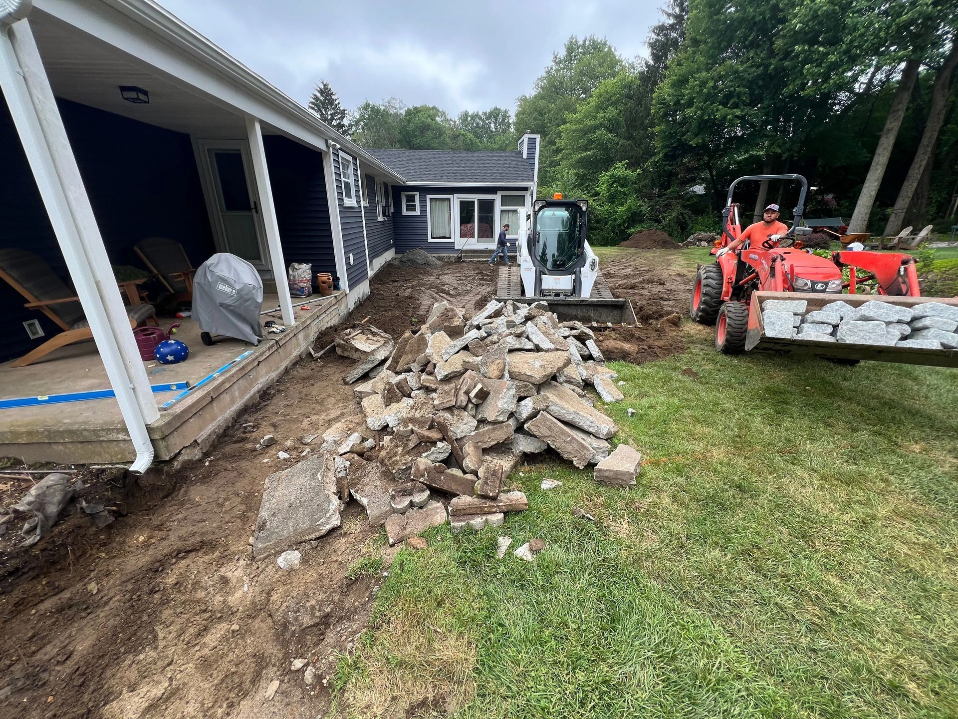 Construction equipment clearing debris beside a house with a covered porch and dirt trench.