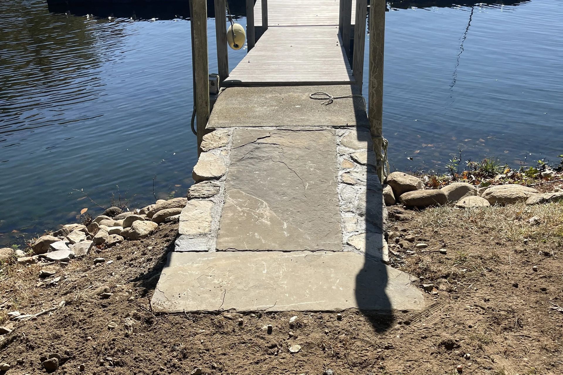 Concrete dock ramp leading to a narrow wooden pier over calm water, with rocks along the shore