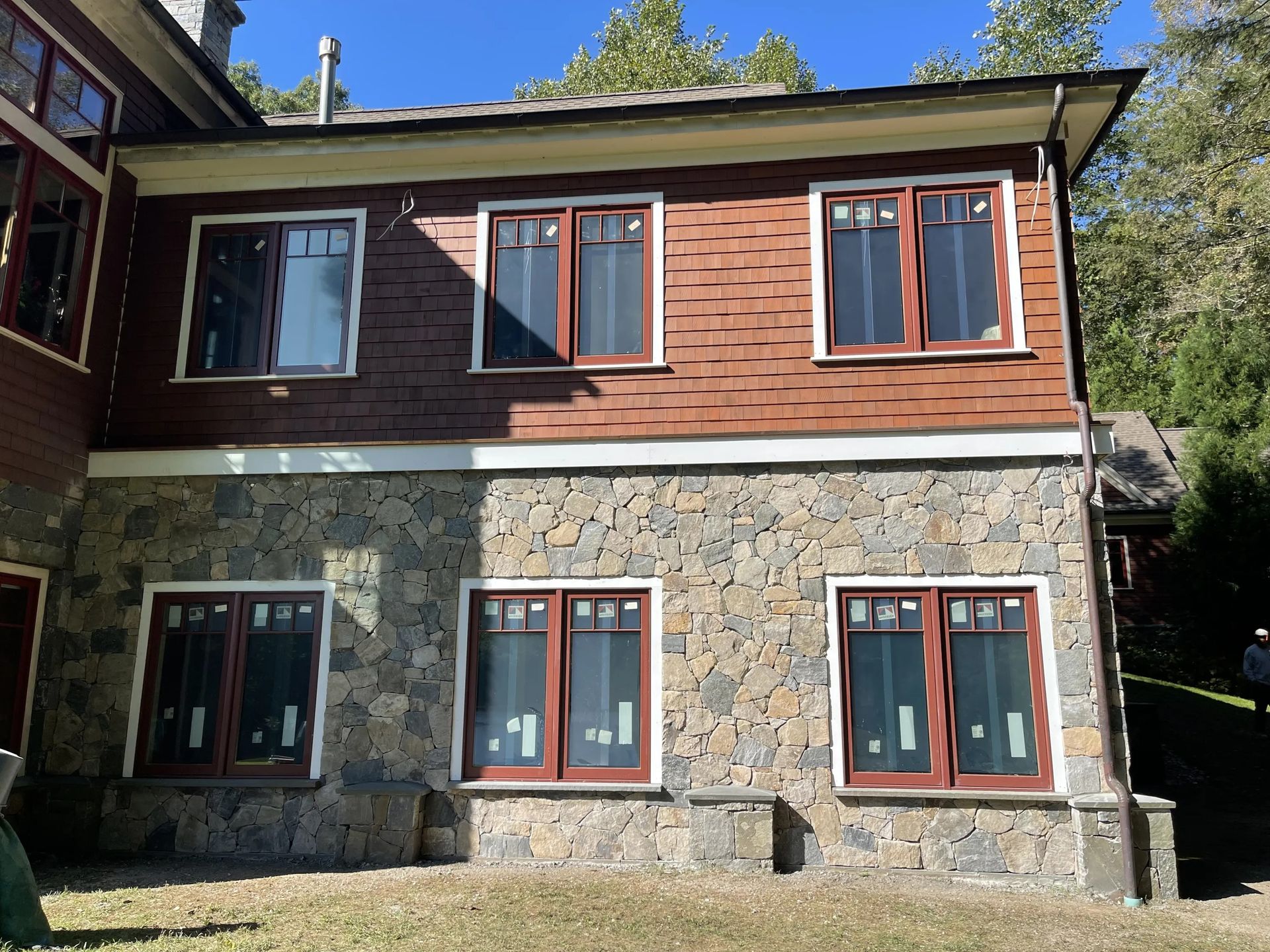 Two-story stone and wood house with red-framed windows in a sunny wooded setting