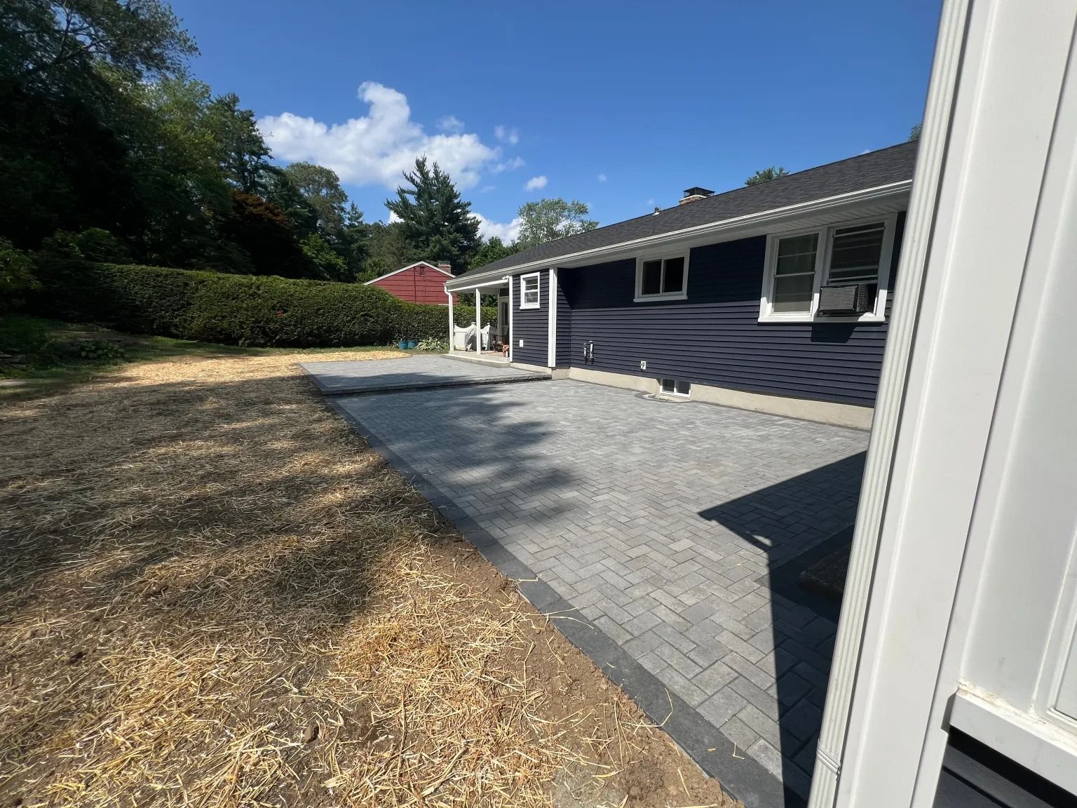 Backyard patio with gray pavers beside a dark blue house, bordered by dry grass and trees