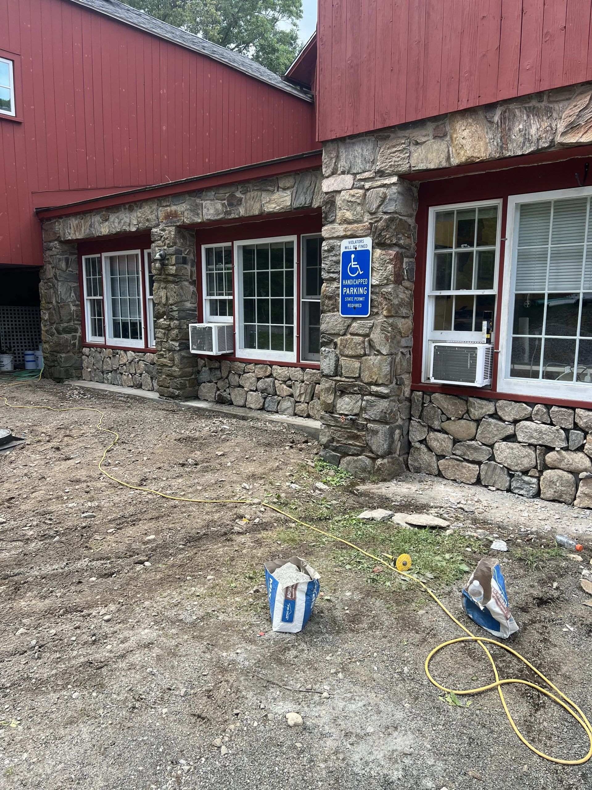 Stone building with red siding, white-framed windows, and a blue sign on a gravel path with hoses and paint cans.