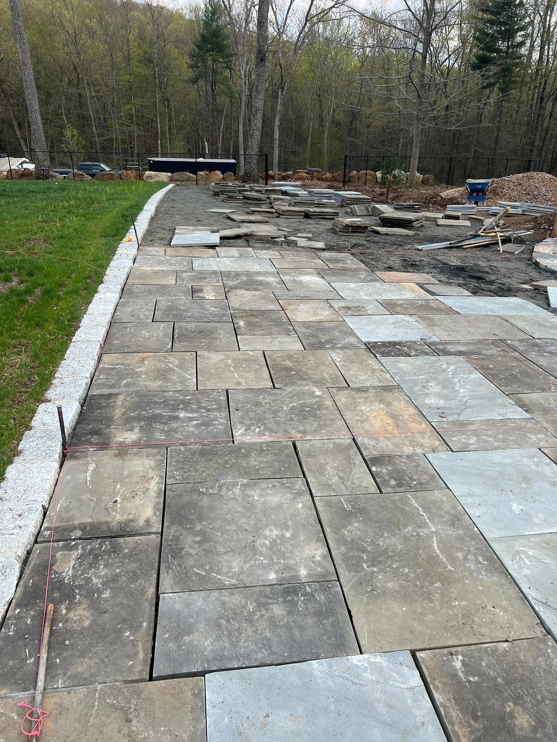 Stone patio walkway beside a grassy lawn, leading past a partially cleared yard with trees and debris.