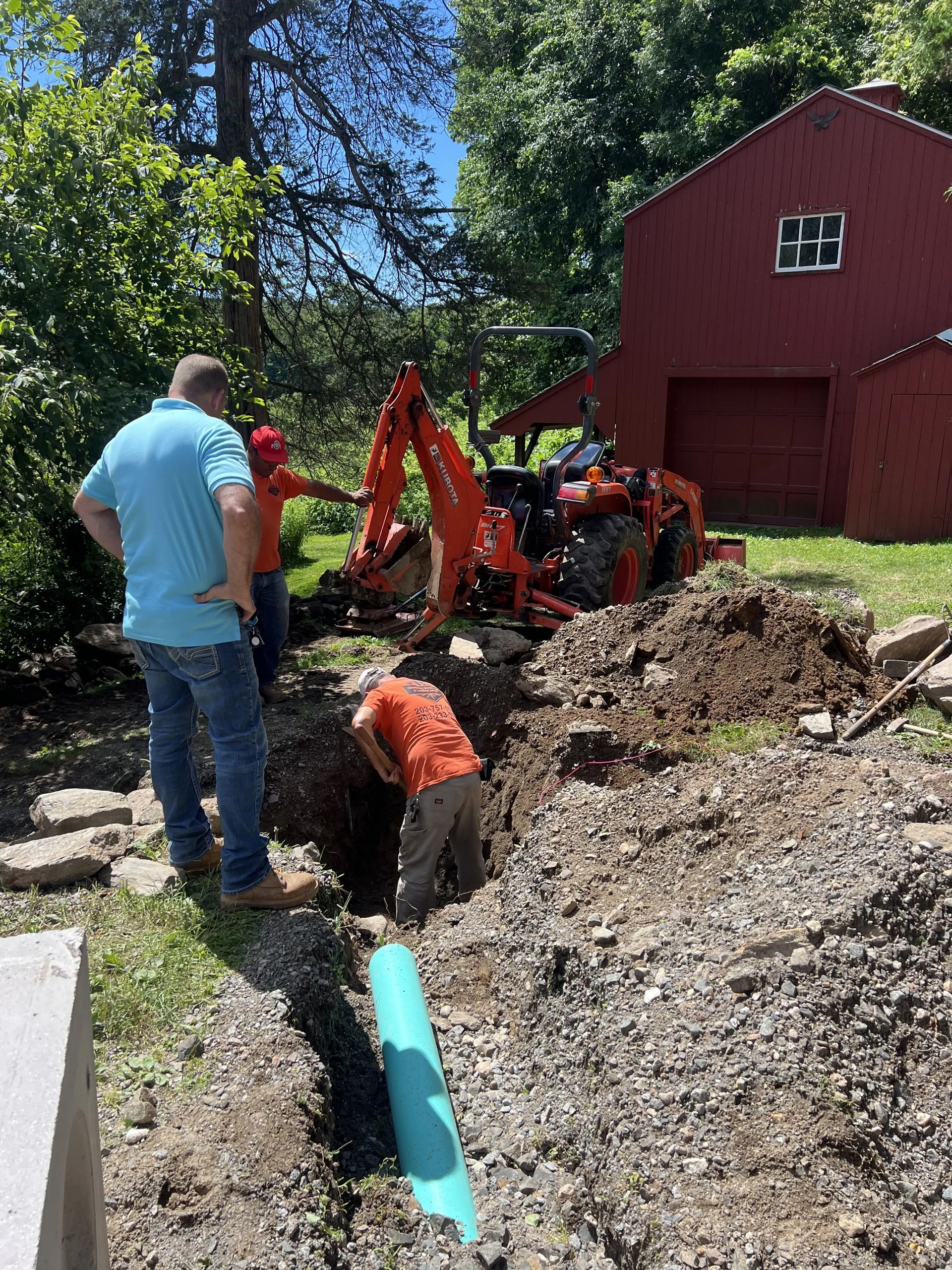 Workers and a backhoe digging a trench beside a red barn in a wooded yard