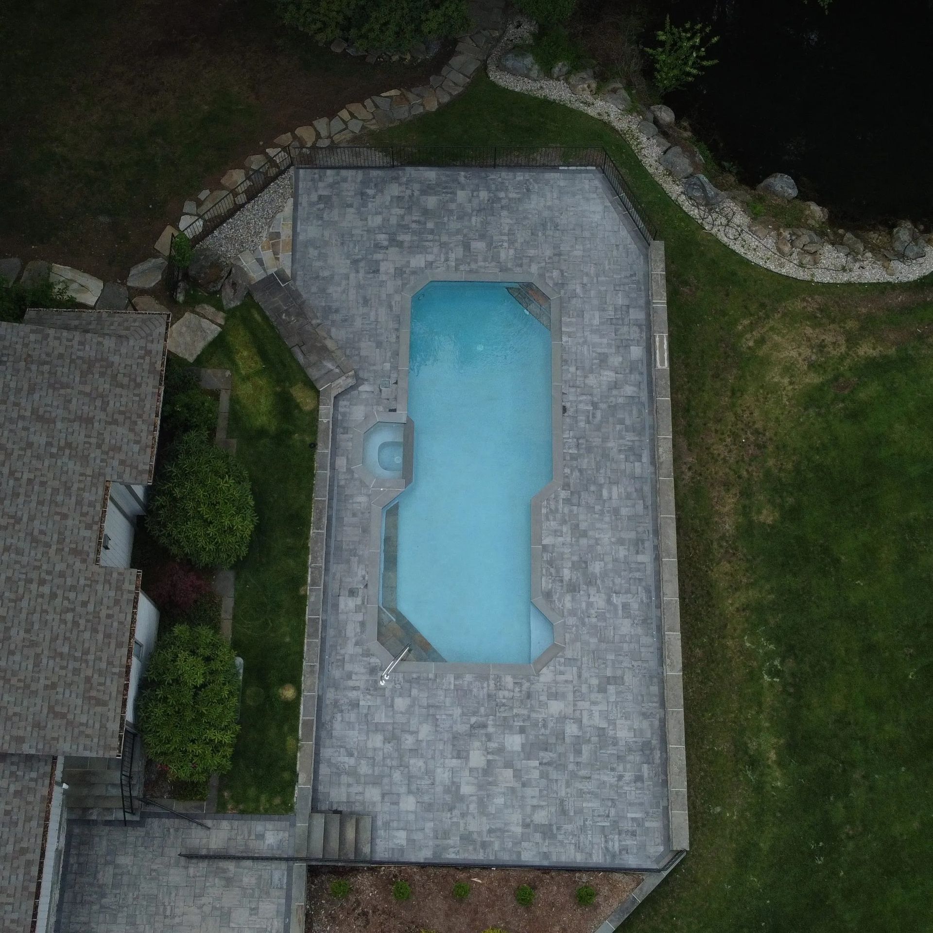 Aerial view of a backyard pool with gray stone patio and surrounding grass and landscaping.