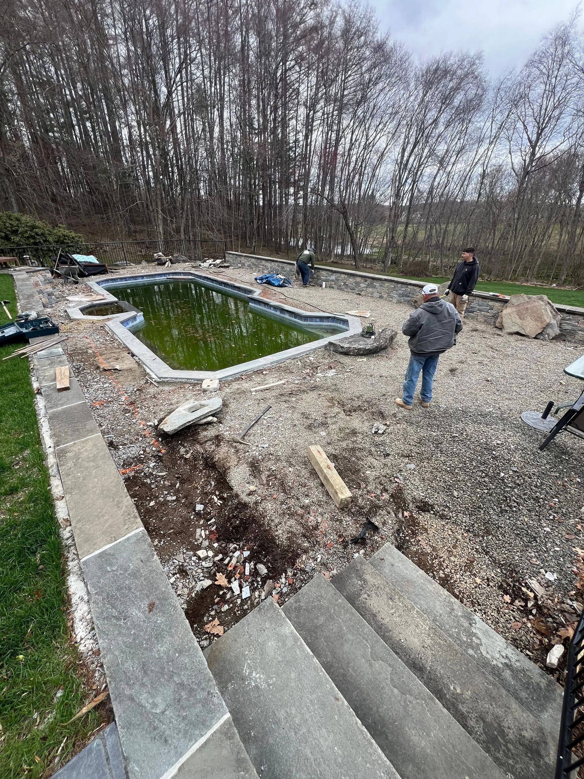 Gravel courtyard with a small green pond, stone steps, and two people near leafless trees