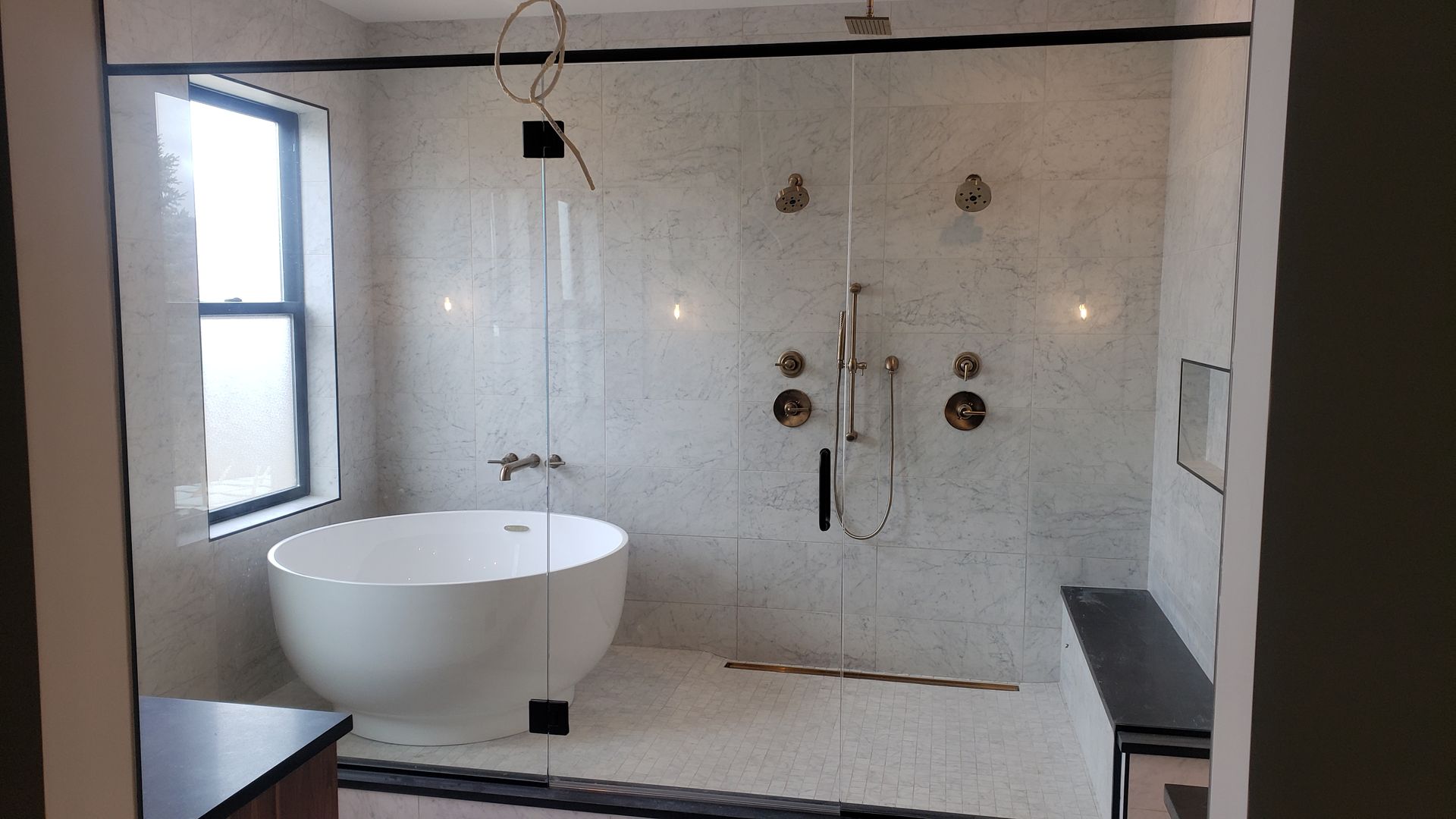 Modern tiled bathroom with a freestanding tub, walk-in shower, window, and dark bench seat.
