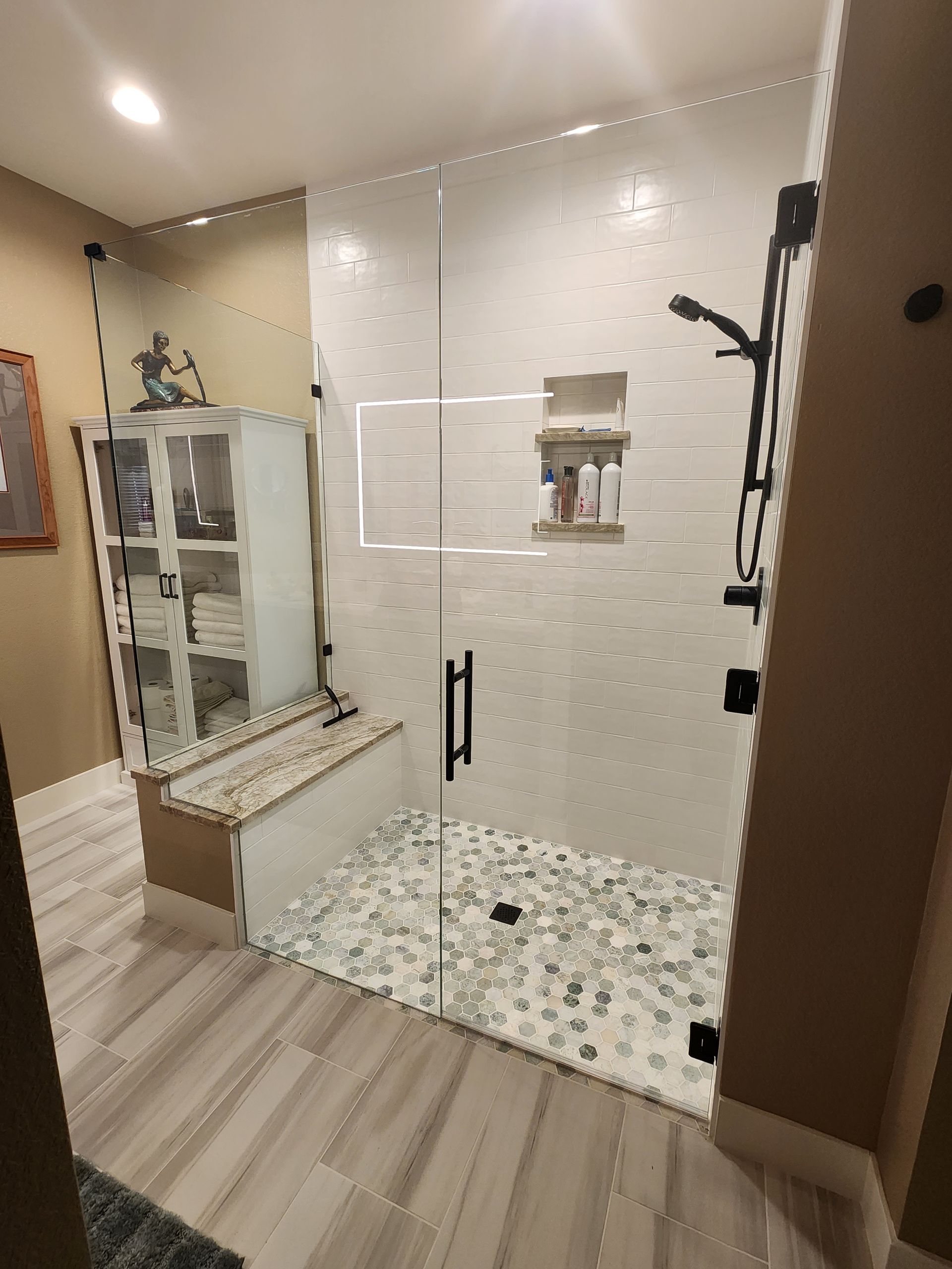 Modern bathroom with white tiled shower, glass door, and mosaic pebble floor beside wood cabinetry.