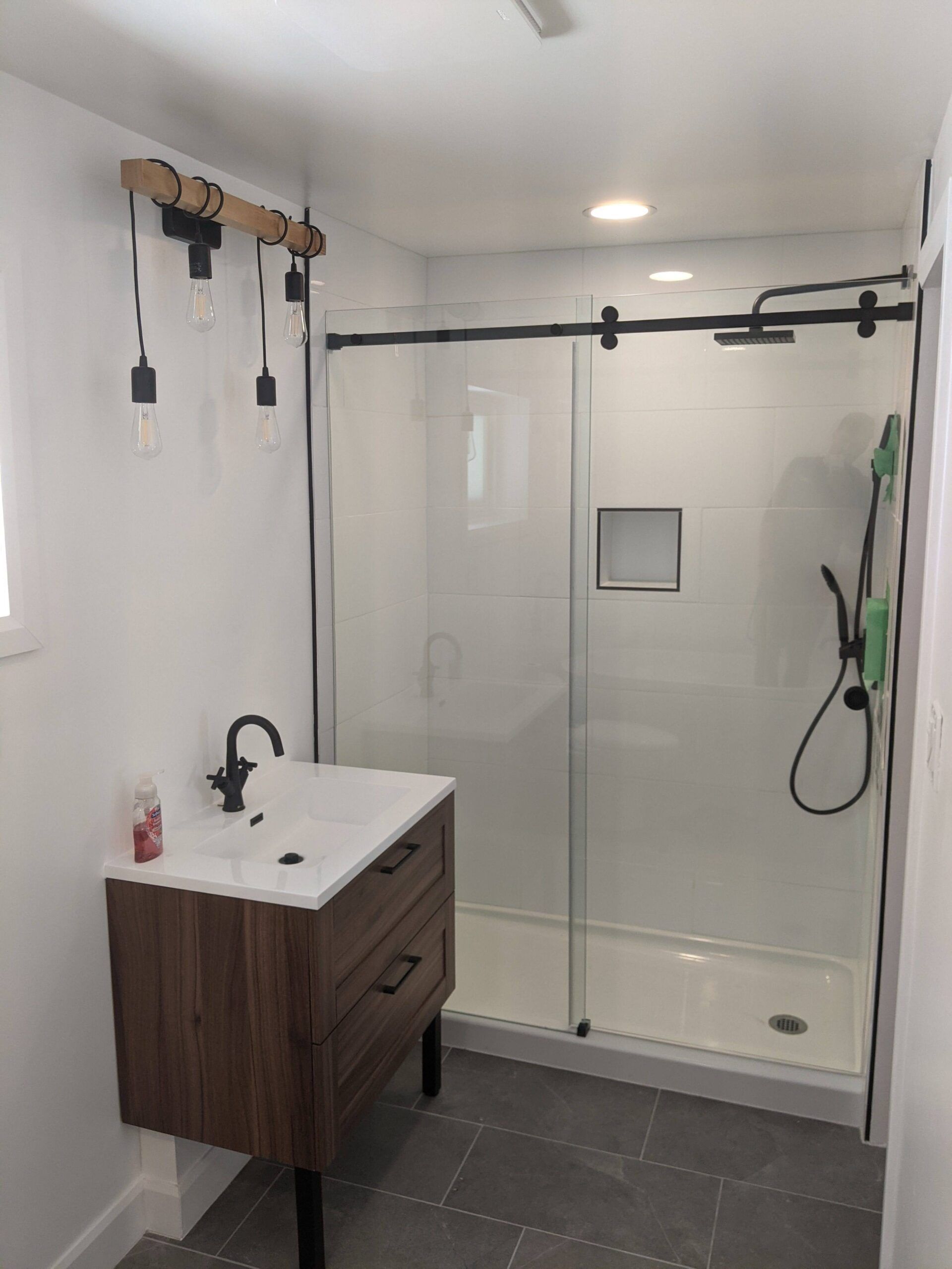 Modern bathroom with glass shower, wood vanity, gray tile floor, and wall-mounted lights