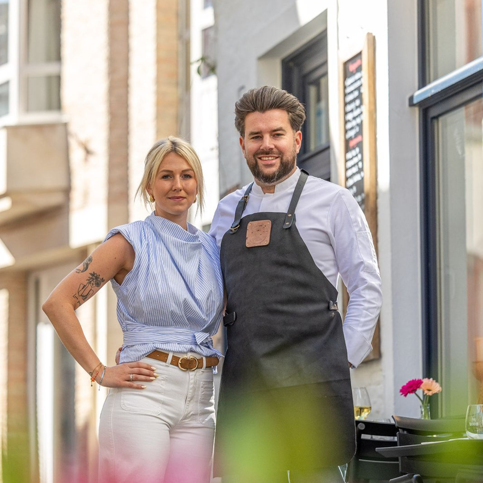 Two smiling people standing outside a shop, one in an apron, with pink flowers blurred in the foreground