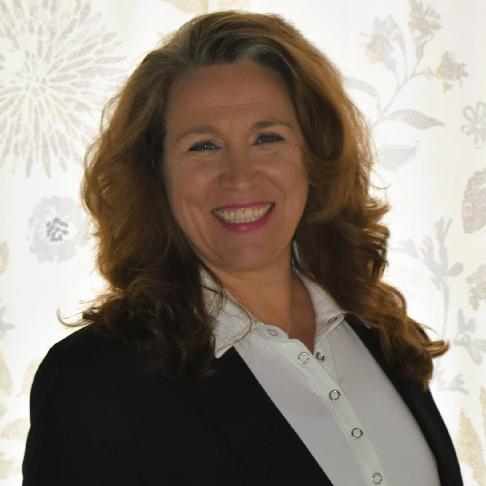 A smiling professional with wavy brown hair wearing a white shirt and dark blazer against a floral backdrop.