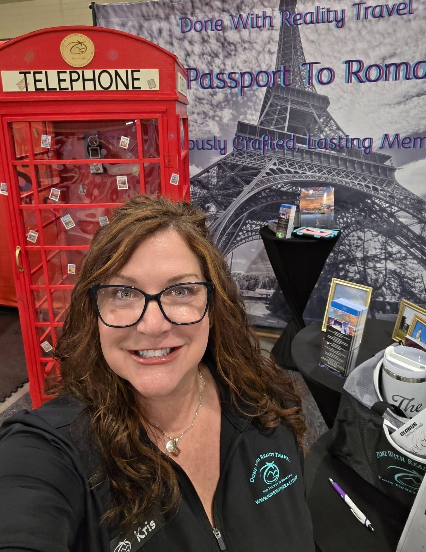 A person with long brown hair wearing glasses and a dark shirt smiles in front of a travel agency booth with an Eiffel Tower.