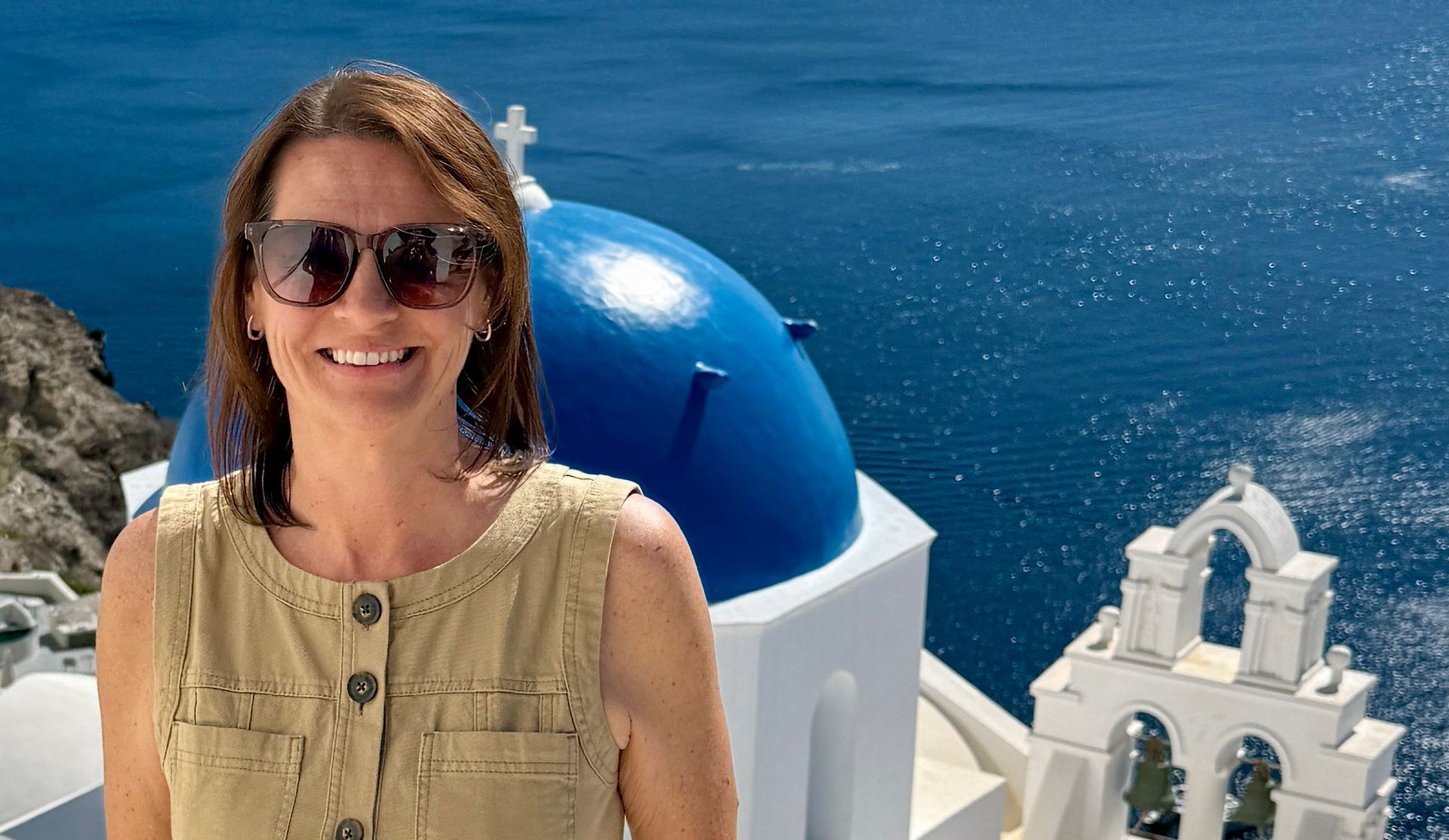A smiling person wears sunglasses and a beige top in front of the iconic blue-domed church and sea of Santorini, Greece.