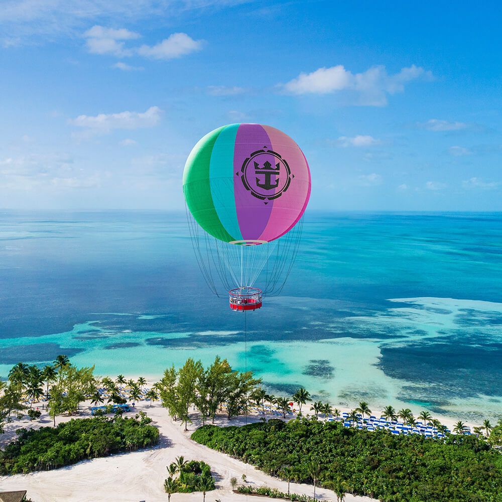 A colorful hot air balloon with a Royal Caribbean logo floats over a tropical beach and turquoise ocean.