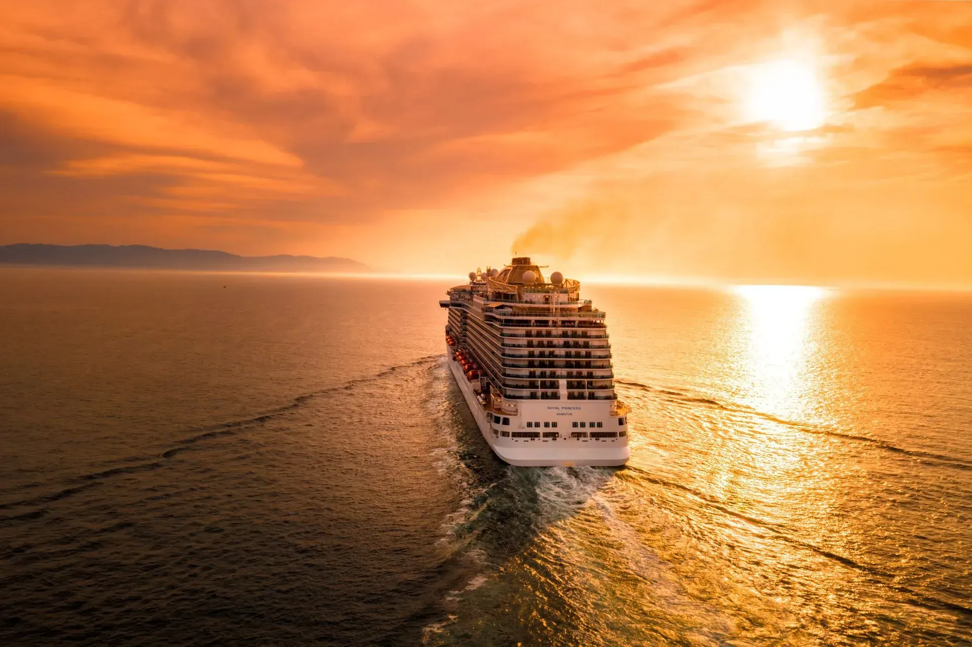 A large cruise ship sails across a calm, reflective ocean during a vibrant orange sunset.