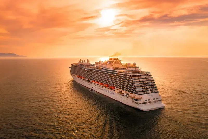 A large cruise ship sails on the ocean during a vibrant orange sunset.