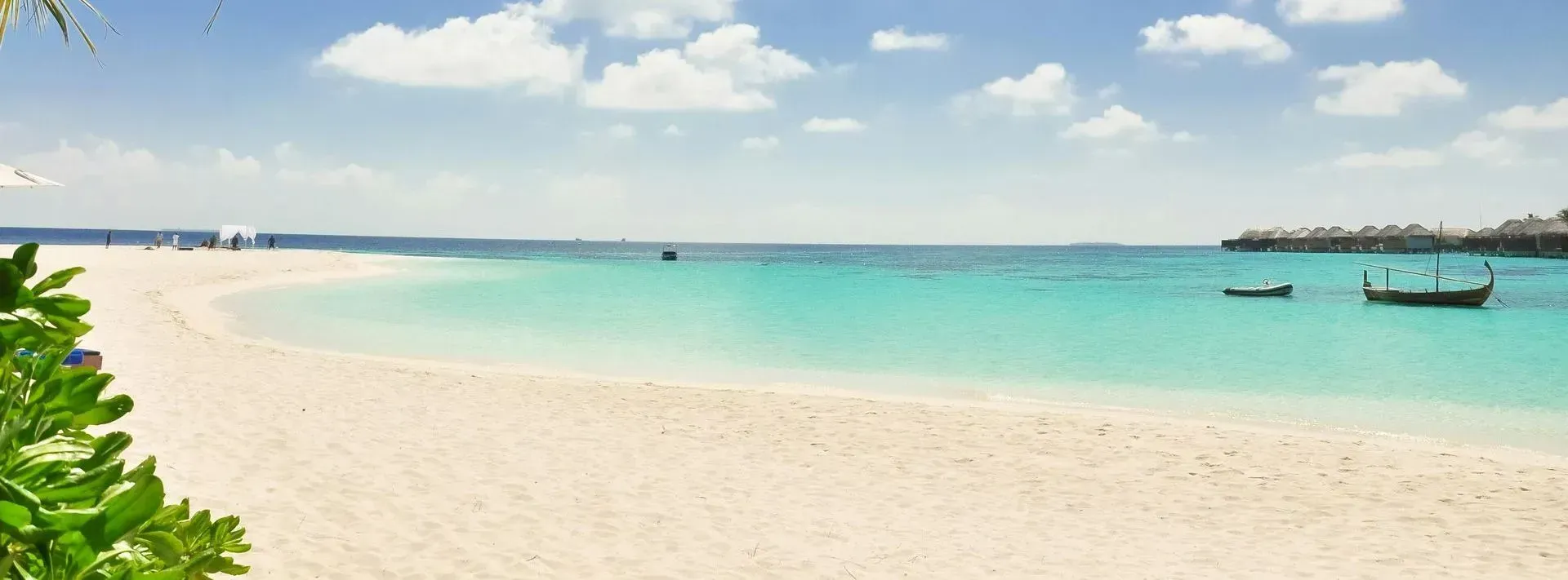 A sandy beach meets turquoise water under a blue sky with white clouds, with small boats anchored in the distance.