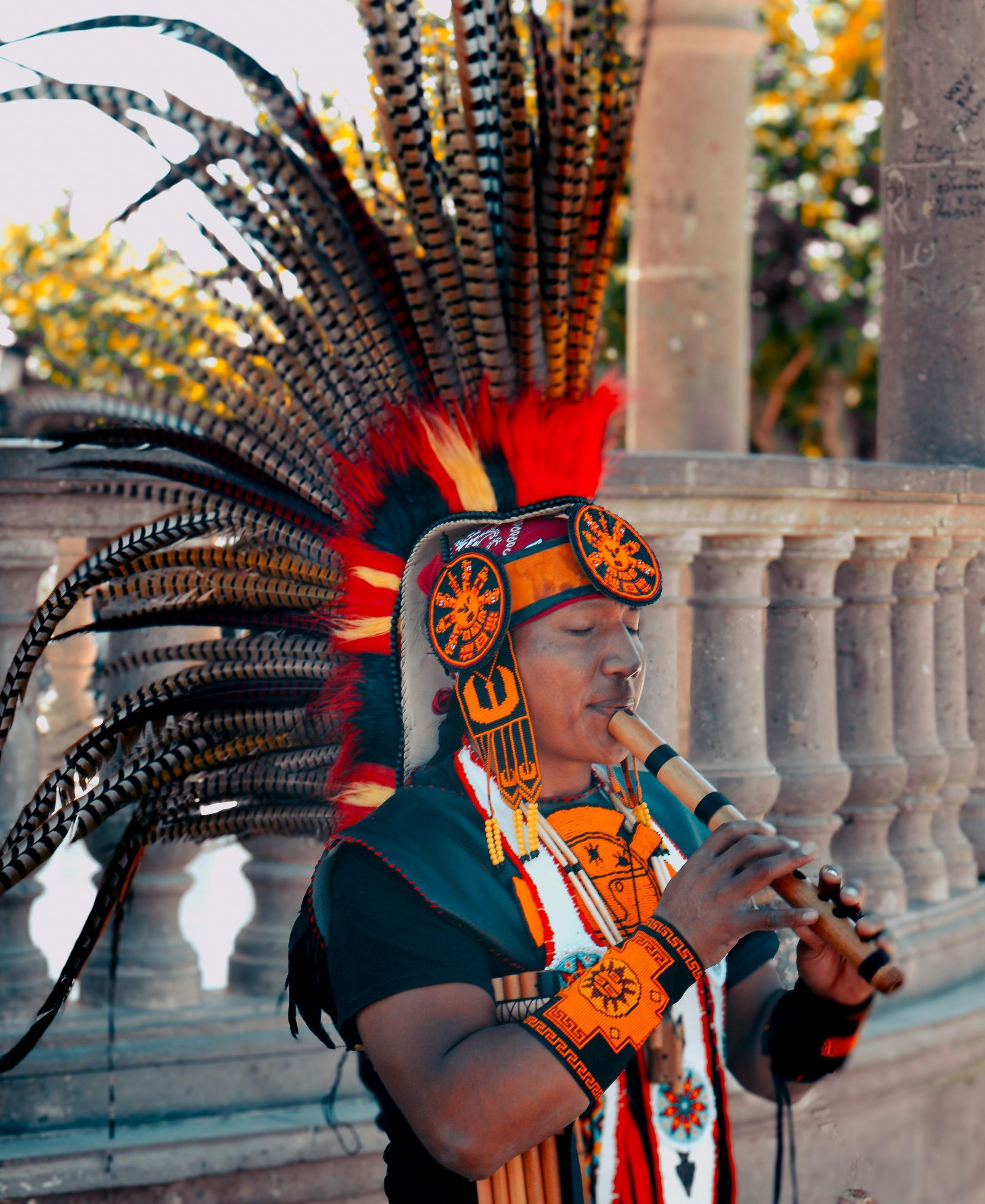 A performer in a vibrant feathered headdress and traditional attire plays a wooden flute against a stone balustrade.