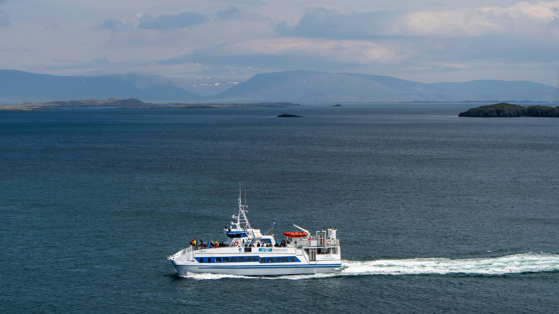 A white passenger ferry cruises across calm, dark blue water toward distant mountains under a cloudy sky.