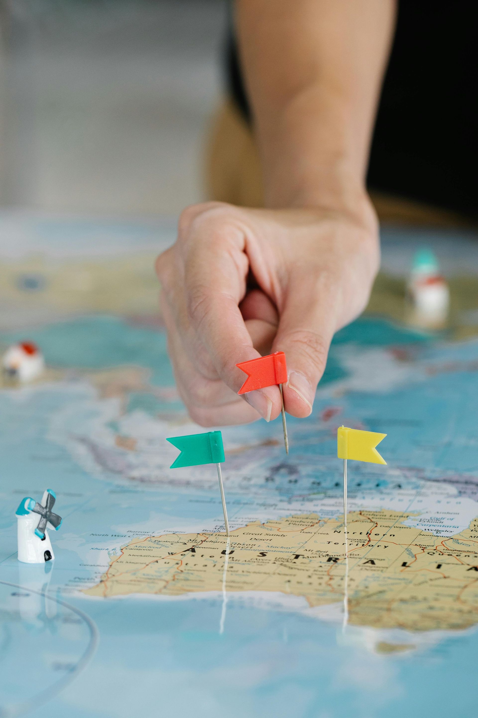 A hand placing a red flag pin on a map of Australia, near green and yellow flag pins and a small model windmill.