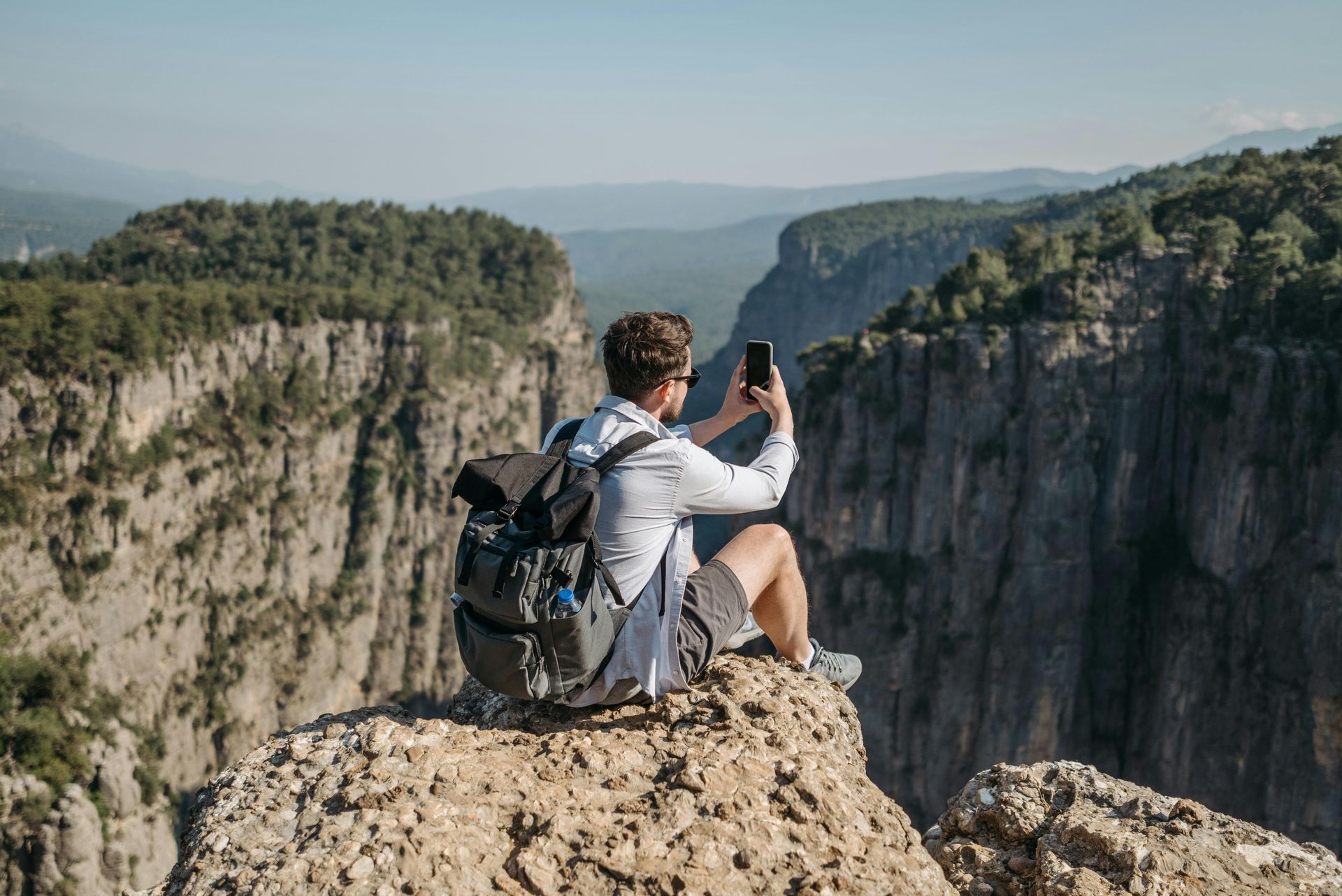 A hiker sits on a rocky cliff edge, using a phone to photograph a deep mountain canyon.