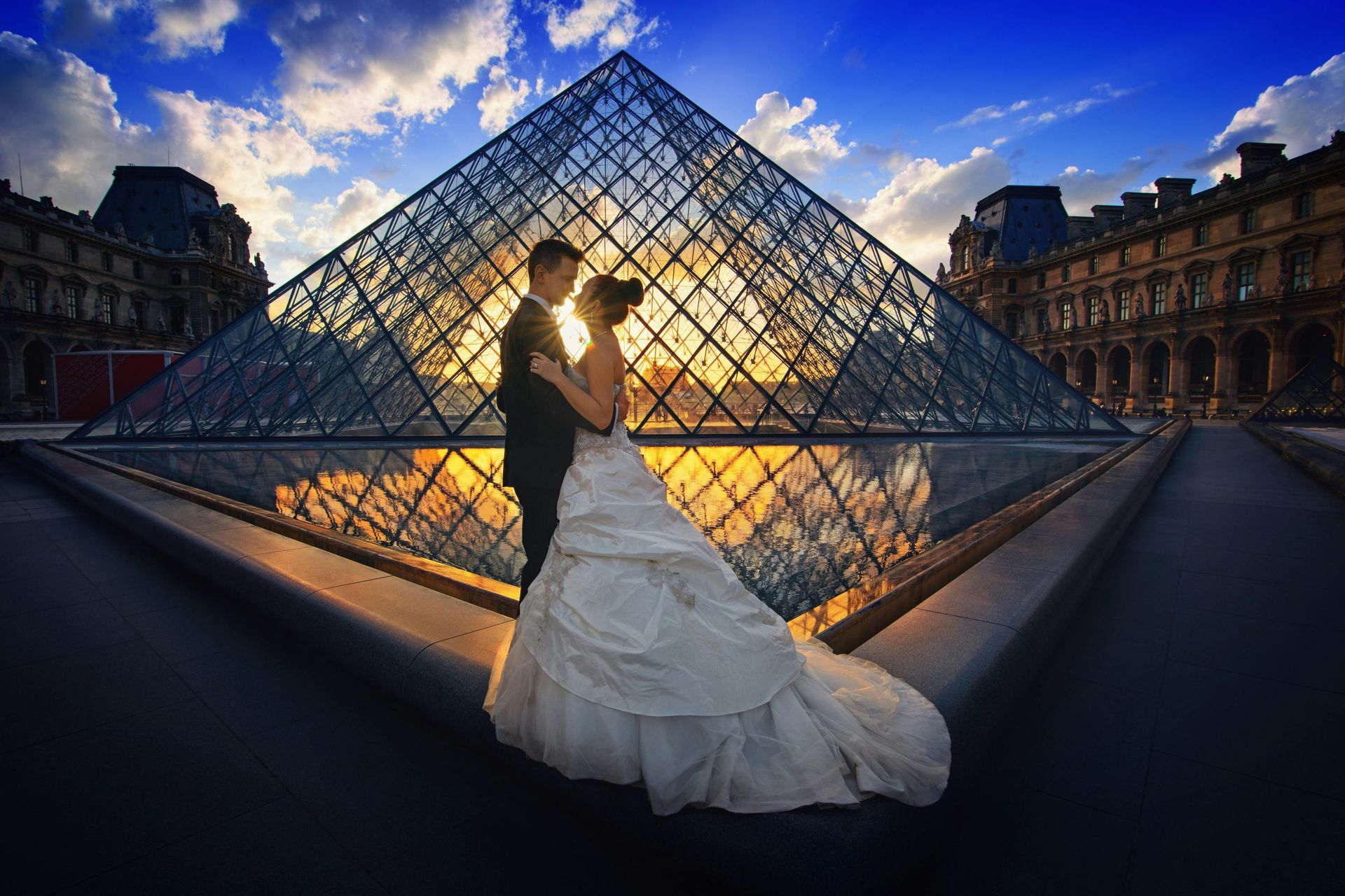 A couple stands in wedding attire before the Louvre Pyramid at sunset, with golden light reflecting off the glass.