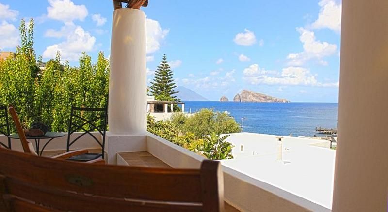 Patio ombreggiato in riva al mare con colonne bianche, sedia in legno, oceano blu e isola rocciosa in lontananza sotto un cielo luminoso