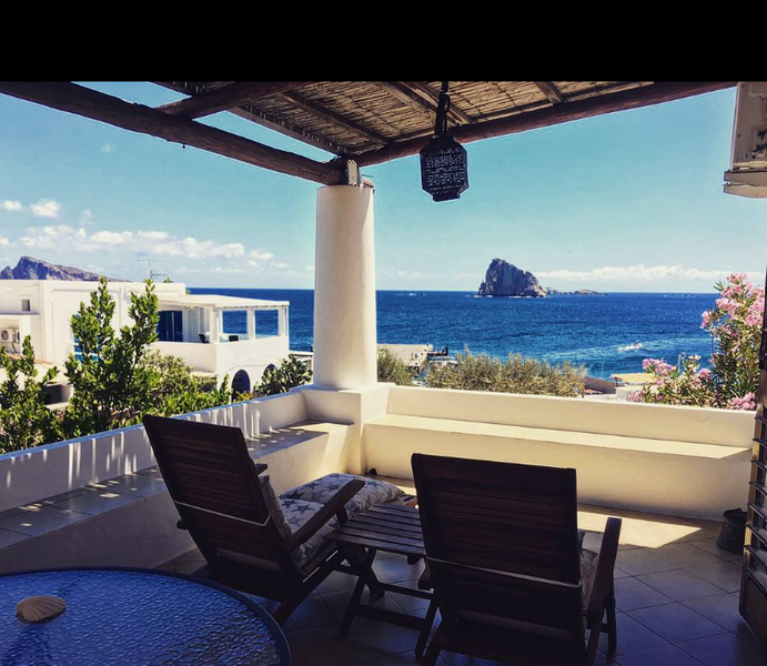 Patio coperto sul mare con lettini, vista sull'oceano, cielo azzurro e un isolotto roccioso in lontananza.
