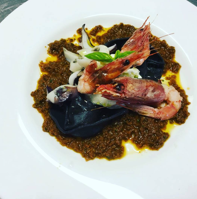 Piatto gourmet di pesce con gamberi e calamari in salsa scura su un piatto bianco.