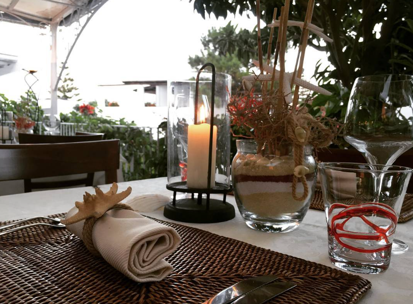 Tavolino da caffè illuminato da candele con tovagliolo arrotolato, bicchiere d'acqua e piante in vaso in un ambiente da giardino.