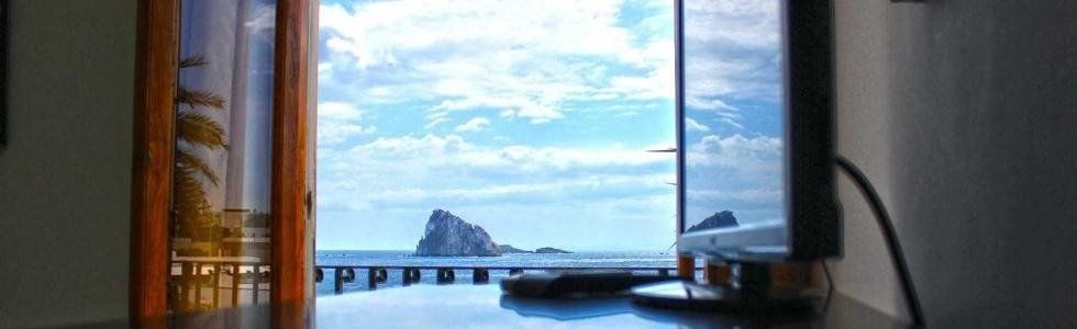 Vista dalla finestra di un molo sul mare con uno scoglio sotto un cielo blu, incorniciata da tende e una bottiglia.