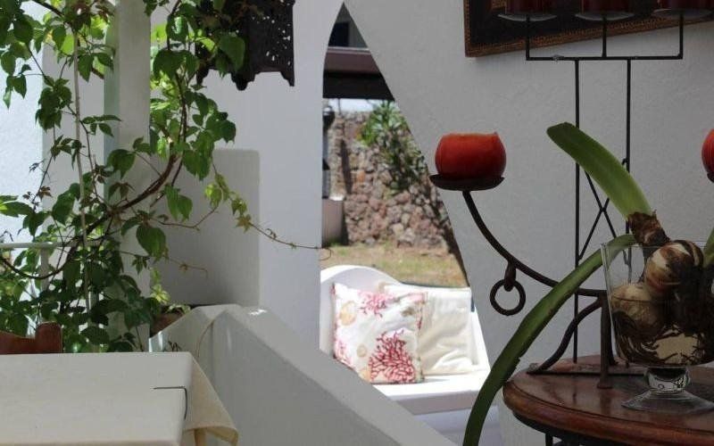 Vista sul giardino del patio con muro bianco, candela rossa, piante in vaso e un angolo con cuscini floreali oltre un arco.
