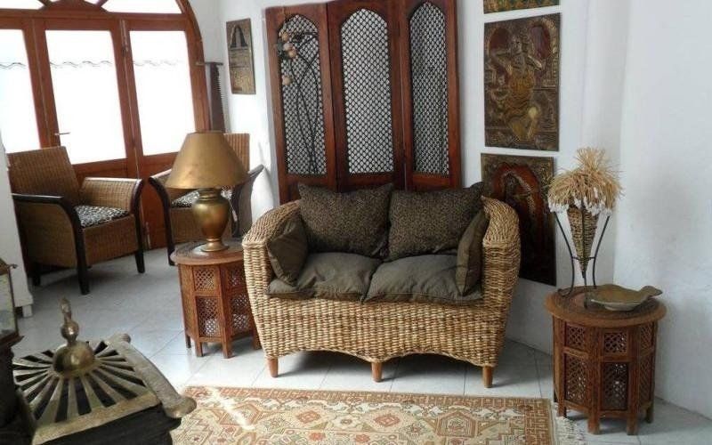 Accogliente divanetto in vimini con cuscini decorati in un luminoso soggiorno, affiancato da tavolini e lampade.