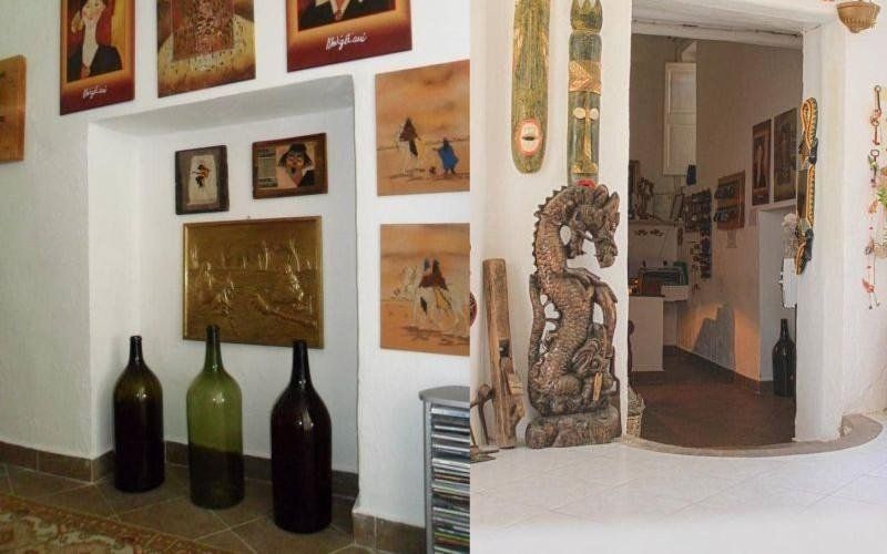 Una stanza simile a una galleria d'arte con opere incorniciate, grandi bottiglie di vino e una scultura decorativa accanto a una porta ad arco.