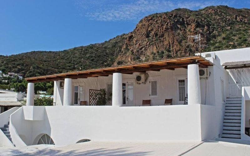 Villa bianca sul mare con terrazza ombreggiata e scala, incastonata tra montagne rocciose sotto un cielo azzurro.