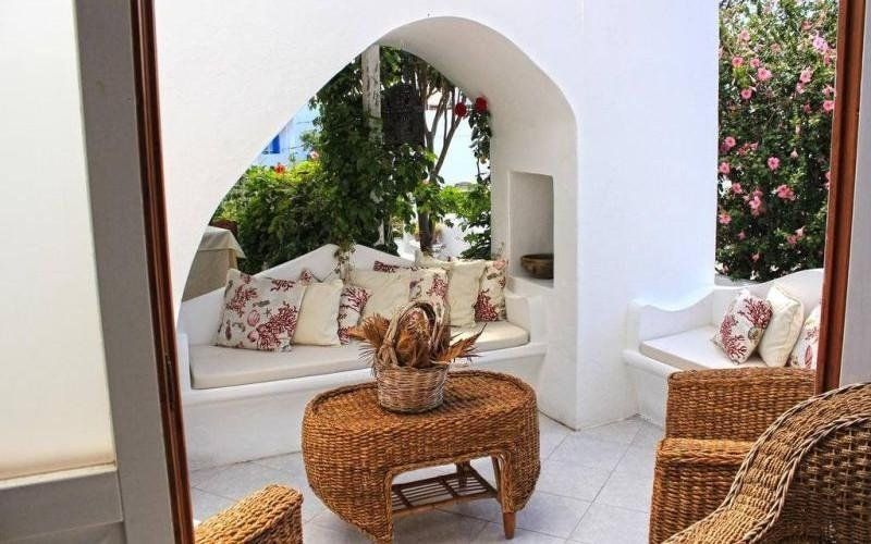 Luminoso patio con sedute ad arco bianche, tavolo in vimini e cuscini floreali, circondato da verde e fiori rosa.