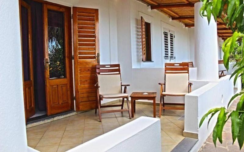 Veranda coperta con sedie in legno, pavimento piastrellato, pareti bianche e una porta in legno accanto a una pianta in vaso.