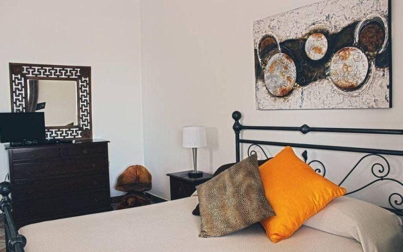 Camera da letto con letto in metallo nero, cuscini arancioni e a fantasia, comò, specchio e quadro astratto alla parete.