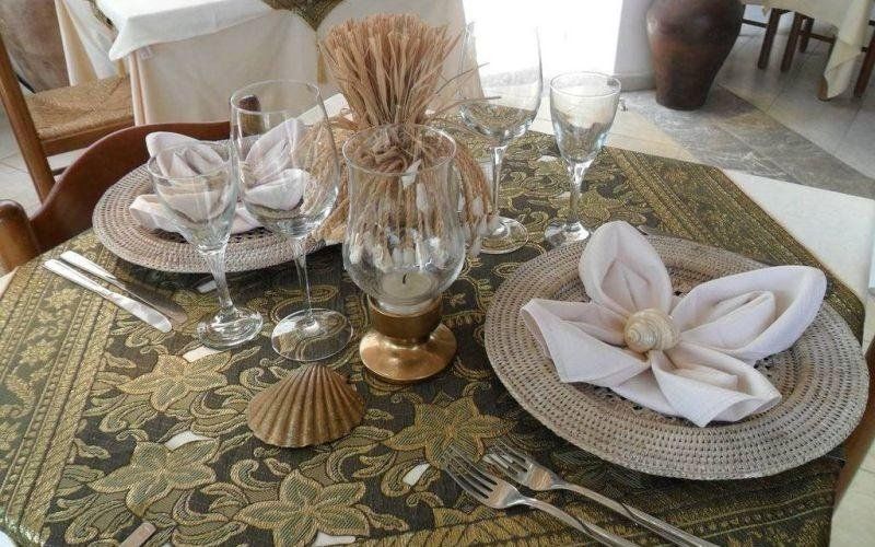 Tavolo da pranzo decorato con tovagliette intrecciate, tovaglioli bianchi, candele e un centrotavola con vaso di vetro.