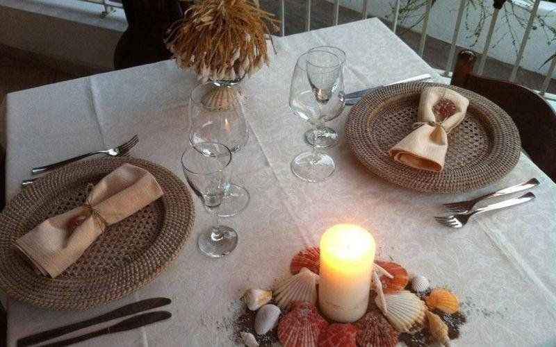 Tavolo da pranzo apparecchiato con tovagliette intrecciate, tovaglioli piegati, bicchieri da vino e centrotavola con candela accesa.