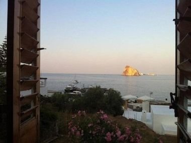 Vista attraverso le persiane aperte di un porticciolo sul mare al tramonto, con un piccolo isolotto roccioso in lontananza.