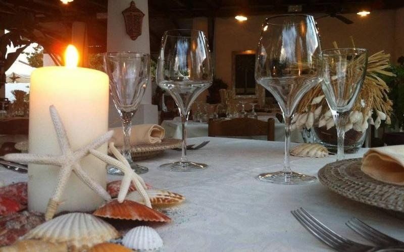Tavolo da pranzo in riva al mare illuminato da candele, con stelle marine, conchiglie, bicchieri da vino e piatti.
