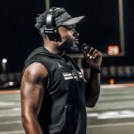 A man wearing headphones and a hat is standing on a football field.
