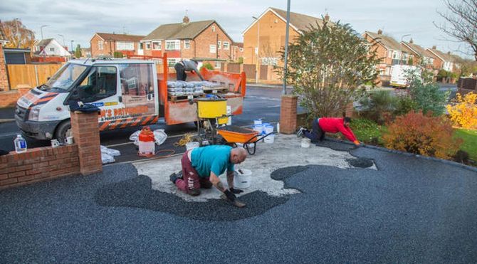 Workers laying black roofing felt on a flat rooftop beside a van and tools in a suburban neighborhood