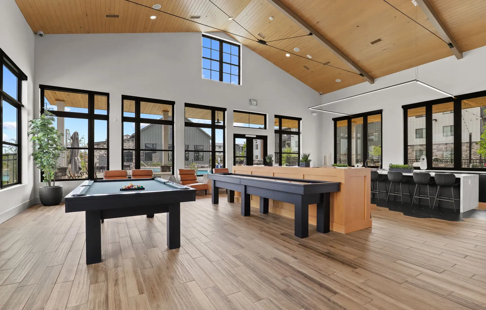 Modern game room with pool table, shuffleboard, lounge seating, and large windows under a vaulted wood ceiling