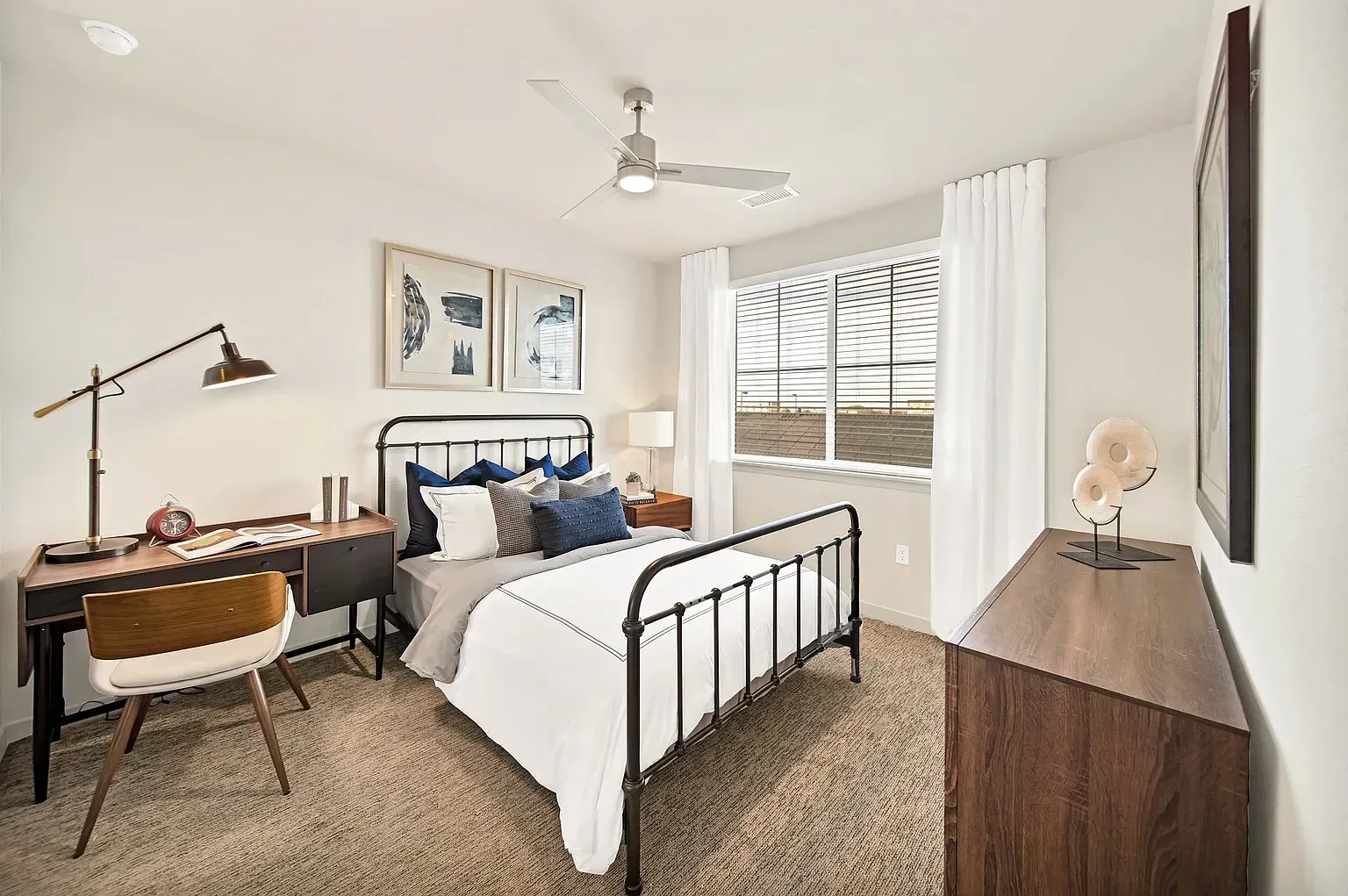 Bright bedroom with white bedding, black metal bed frame, desk, dresser, and window blinds