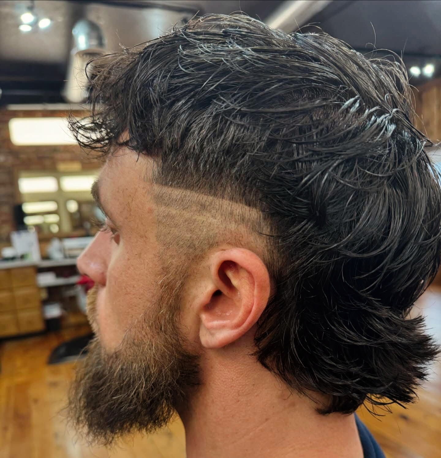 Man with a mullet hairstyle and beard, side profile view in a barbershop.