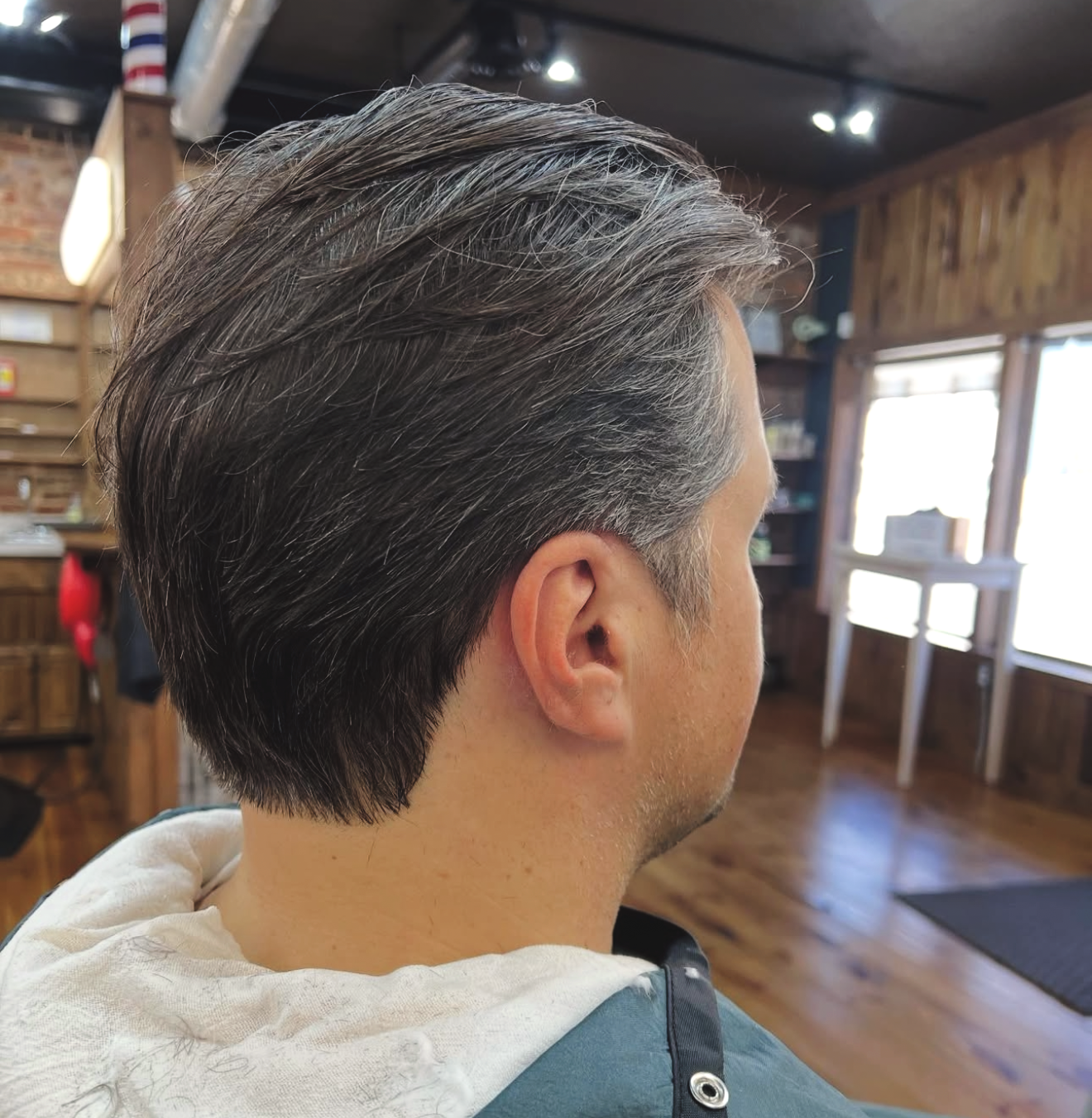 Man in a barber's chair with a fresh haircut; medium length dark hair; in a barbershop with wooden floors.