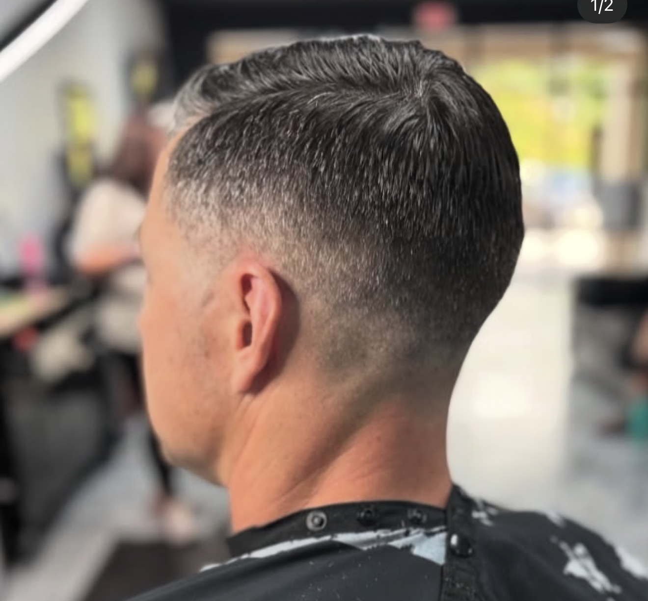 Man's hair styled with a fade cut; dark gray hair, short sides, light skin, barber shop background.