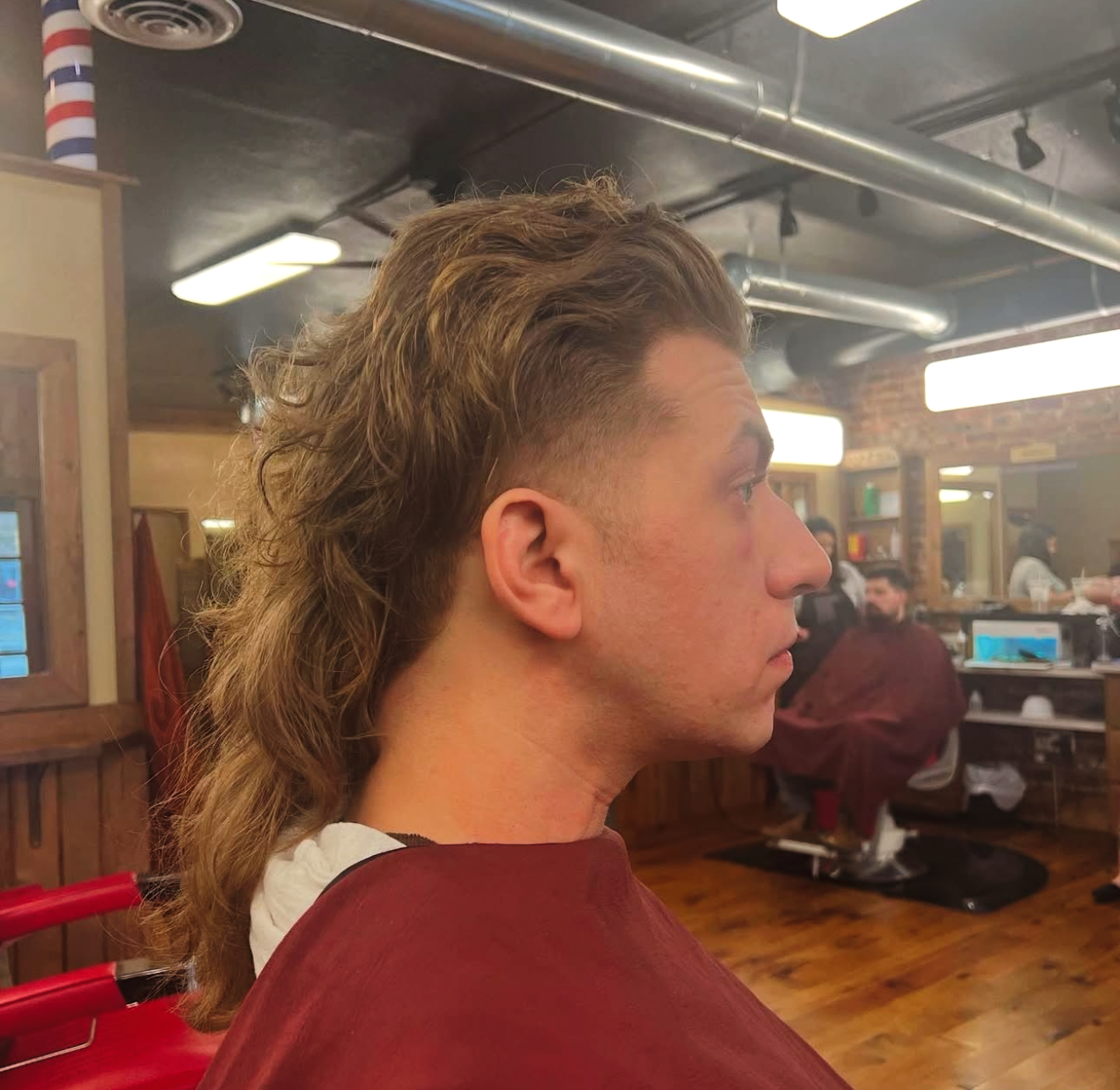 Man with a mullet haircut at a barbershop, side profile view. Light brown hair, reddish barber cape.