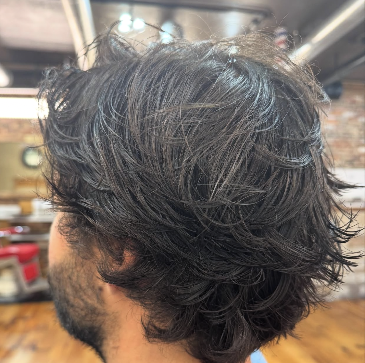 Man with dark, messy hair at a barber shop; side profile view with a dark beard.